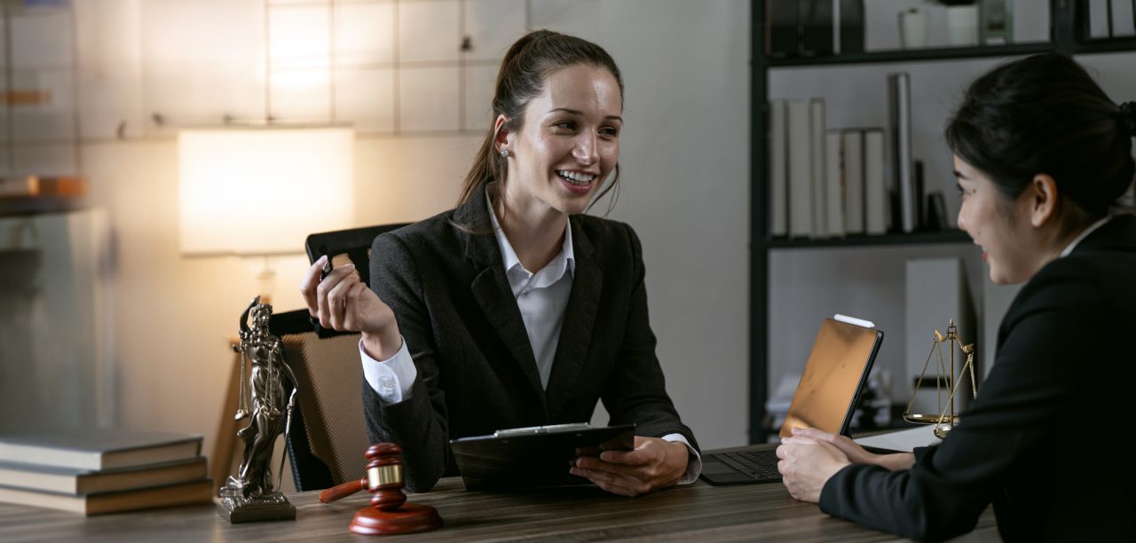 Two women in business attire sit across from each other at a desk in a law office, smiling during a conversation. Legal symbols like a gavel and Lady Justice statue are visible, highlighting Mobile Notary Services New Jersey.