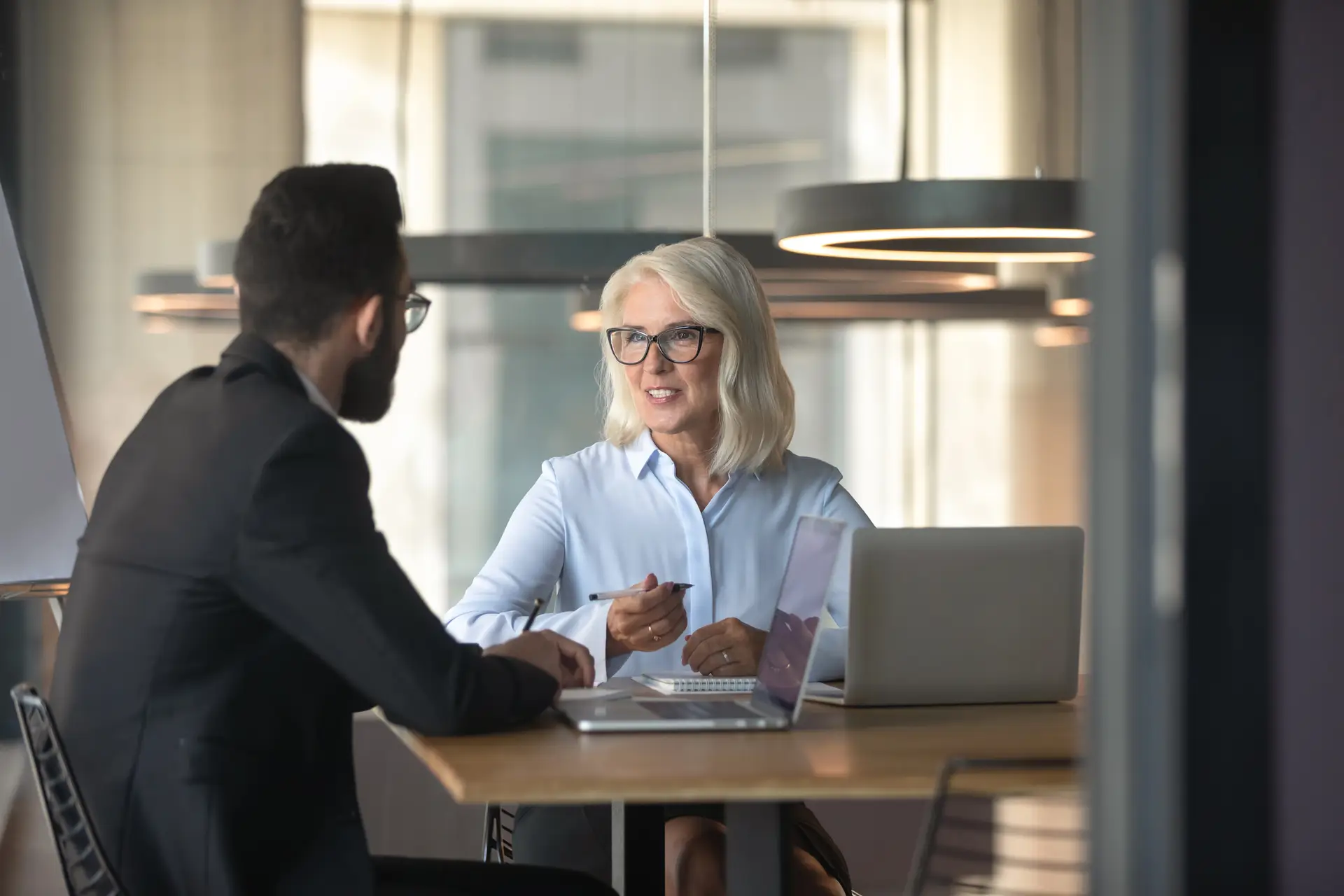 An older woman with gray hair and glasses discusses Legal Document Notarization New Jersey with a bearded man in a suit across a desk. They are in a modern office with laptops and large circular lights overhead.