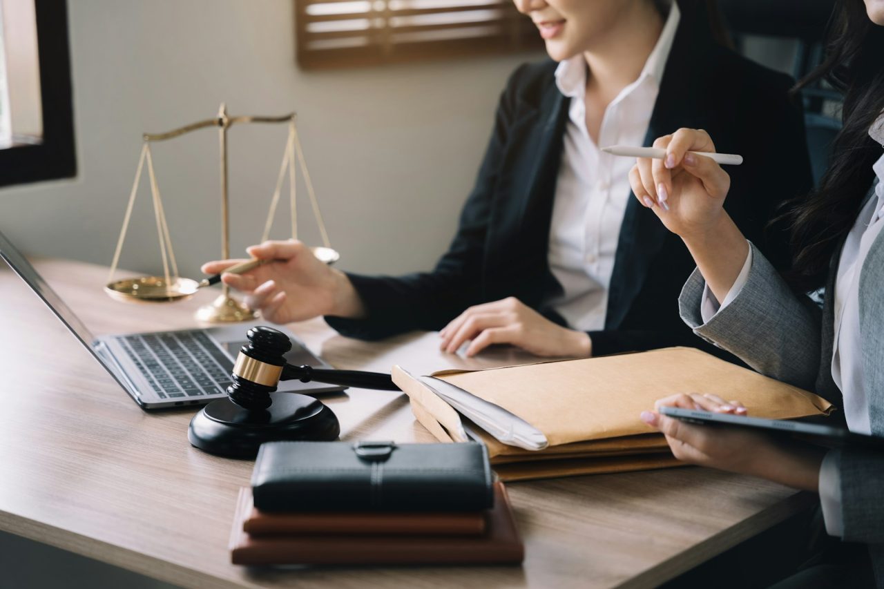 Two professionals in business attire sit at a desk with a laptop, legal documents, a gavel, a balance scale, and a notebook, suggesting a Legal Document Notarization New Jersey meeting in progress.
